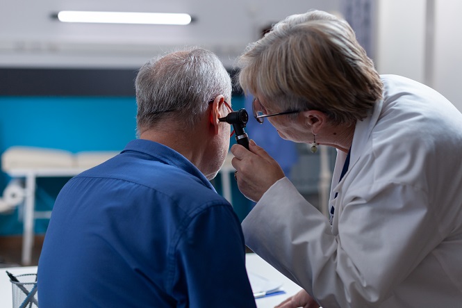 A imagem mostra uma médica examinando o ouvido de um paciente. A médica está vestida com um jaleco branco e usa um equipamento, possivelmente um otoscópio, para inspecionar o ouvido do paciente, que está sentado de costas. A cena parece ocorrer em um ambiente clínico ou consultório médico.