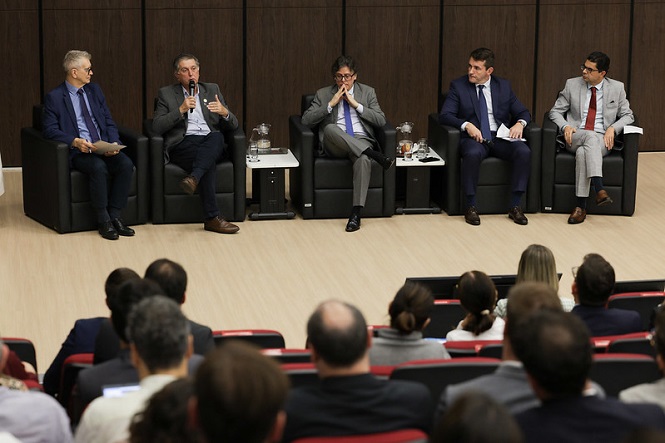 A imagem mostra um homem de terno e gravata, usando óculos, falando em um microfone em um púlpito. No fundo, há uma tela com o logotipo e texto que diz "Seminário Regional da Magistratura Catarinense". O homem parece estar fazendo uma apresentação ou discurso no evento.