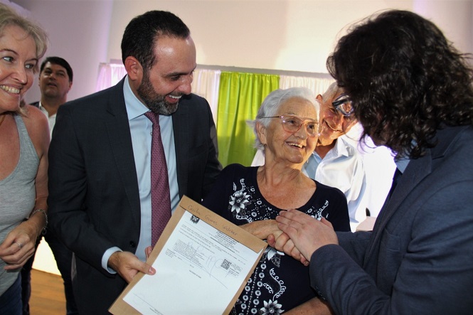 A imagem mostra um grupo de pessoas sorridentes em um evento. Um homem está entregando um documento para uma senhora idosa, que parece estar feliz. Há outras pessoas ao redor, vestidas de maneira formal, incluindo outro homem em terno e uma mulher à esquerda. O ambiente é coberto com uma cortina branca e verde ao fundo.