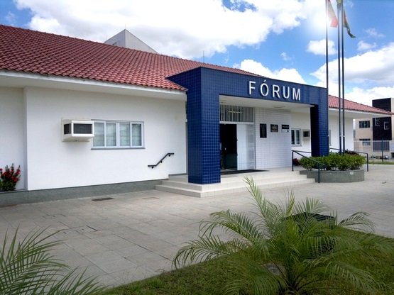 A imagem mostra a fachada de um edifício identificado como "FÓRUM". O prédio tem paredes brancas e um telhado inclinado com telhas vermelhas. Na entrada, há azulejos azuis ao redor da porta principal. Várias bandeiras estão hasteadas em mastros perto da entrada. O local tem algumas plantas e é rodeado por uma área pavimentada. O céu está parcialmente nublado com algumas nuvens brancas.