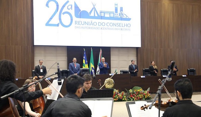 A imagem mostra a 26ª Reunião do Conselho das Autoridades Centrais Brasileiras. Há uma mesa com várias pessoas em pé, possivelmente autoridades, e bandeiras ao fundo. Na parte inferior da imagem, há músicos tocando instrumentos de orquestra. O ambiente é formal, com um painel grande atrás mostrando o nome do evento.
