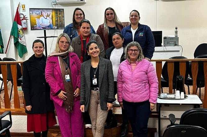 A imagem mostra um grupo de pessoas posando para uma foto em um ambiente que parece ser uma sala de conferências ou reunião. Há nove pessoas ao todo, seis delas em pé na parte de trás e três na frente. Algumas das pessoas estão usando crachás, sugerindo que são profissionais no local de trabalho. No fundo, há uma bandeira ao lado de uma tela que exibe uma imagem. Ao lado esquerdo, há uma mesa com uma bandeja e uma garrafa térmica. O ambiente parece formal e organizado.