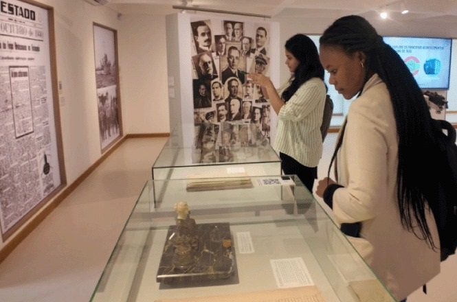 Na imagem, há duas mulheres, uma negra em primeiro plano e uma mulher branca em segundo plano,  em um ambiente de museu, demonstrando interesse nas peças em exibição. Elas olham para objetos dentro de vitrines de vidro. Ao fundo, há várias fotografias em uma parede e um grande painel de jornal antigo exibido na parede lateral. As mulheres parecem estar observando e possivelmente fotografando as exposições. O museu parece ter um tema histórico, como indicado pelos elementos presentes.