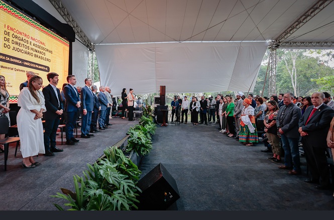 A imagem mostra um evento solene em um palco coberto, onde diversas pessoas estão de pé, vestidas formalmente. Há uma fila de pessoas no palco, incluindo homens e mulheres usando trajes formais, como ternos e roupas sociais. Alguns deles parecem estar fazendo discursos ou apresentações. O público está de pé em frente ao palco, e há uma decoração com plantas. No fundo, a faixa indica que o evento é o "I Encontro Nacional de Direitos Humanos do Judiciário Brasileiro". A atmosfera é de formalidade e respeito.
