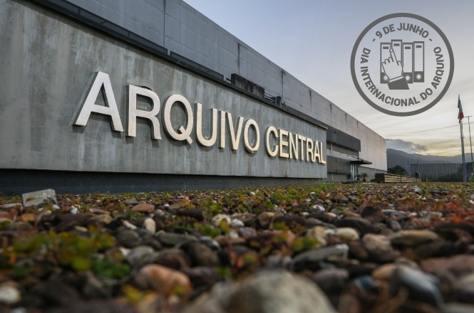 A imagem mostra um prédio com uma fachada onde está escrito "ARQUIVO CENTRAL" em letras grandes. Em primeiro plano, há um solo coberto de pequenas pedras e algumas plantas. No canto superior direito, há um selo cinza que diz "9 DE JUNHO - DIA INTERNACIONAL DO ARQUIVO" com um desenho estilizado de arquivos. O céu ao fundo está parcialmente nublado.