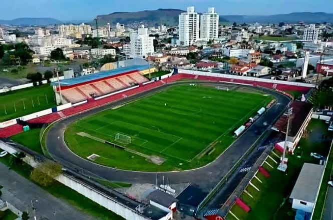 A imagem mostra um estádio de futebol, visto de cima. O campo é gramado e cercado por uma pista de atletismo. Há arquibancadas de cimento em ambos os lados do campo. Ao fundo, é possível ver vários prédios e uma área urbana, com casas e edifícios. As cores predominantes nas arquibancadas são vermelho e branco. O ambiente em volta parece uma cidade com algumas colinas ao fundo.