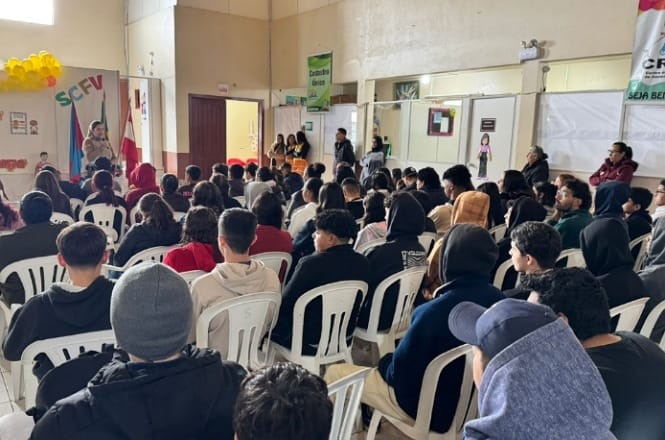 A imagem mostra uma sala cheia de pessoas sentadas em cadeiras de plástico branco, aparentemente assistindo a uma apresentação ou palestra. Há uma pessoa em pé na frente   da plateia, falando ou apresentando algo. O ambiente parece ser um espaço comunitário ou escolar, com algumas bandeiras e cartazes na parede. As pessoas estão vestidas de maneira casual e prestam atenção no que está sendo apresentado. Há ainda alguns elementos decorativos, como balões.