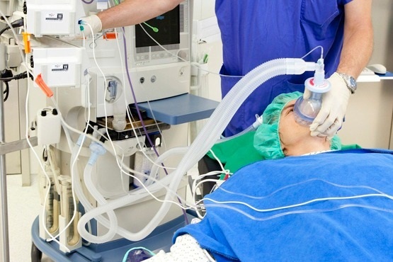 A imagem mostra uma pessoa deitada em uma maca, coberta com um lençol azul. A pessoa está sendo atendida em um ambiente que parece ser uma sala de cirurgia, pois há um profissional de saúde ao lado dela, vestindo roupas médicas azuis e segurando uma máscara de oxigênio sobre o rosto do paciente. Ao lado, há máquinas médicas com vários cabos e tubos conectados. O ambiente e os equipamentos sugerem uma situação de cuidados médicos intensivos, possivelmente relacionada à anestesia ou respiração assistida.