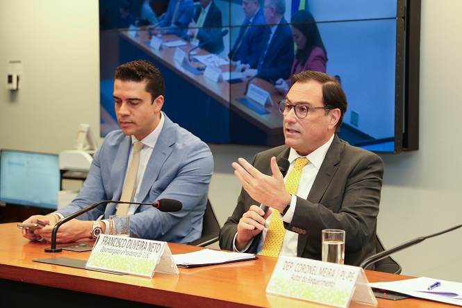 A imagem mostra dois homens em uma reunião formal, provavelmente em um ambiente de conferência ou audiência. Ambos estão sentados atrás de uma mesa. O homem à esquerda veste um terno azul com gravata amarela e segura um papel. O homem à direita está de terno escuro, gravata amarela e parece estar falando, gesticulando com a mão. Há microfones sobre a mesa e placas de identificação na frente deles. No fundo, uma grande tela exibe a imagem de outras pessoas em uma sala de reuniões.