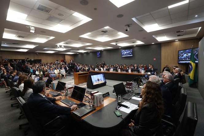 A imagem mostra uma sala de conferência ou reunião cheia de pessoas. Há uma mesa central grande com várias pessoas sentadas ao redor, muitas usando laptops e materiais de leitura. A bandeira do Brasil está visível ao fundo. A sala está equipada com monitores nas paredes que exibem provavelmente conteúdo relacionado à reunião. Há muitas pessoas sentadas no que parece ser uma área de audiência, assistindo ao que está acontecendo na mesa central. O ambiente sugere que é um evento formal ou oficial.