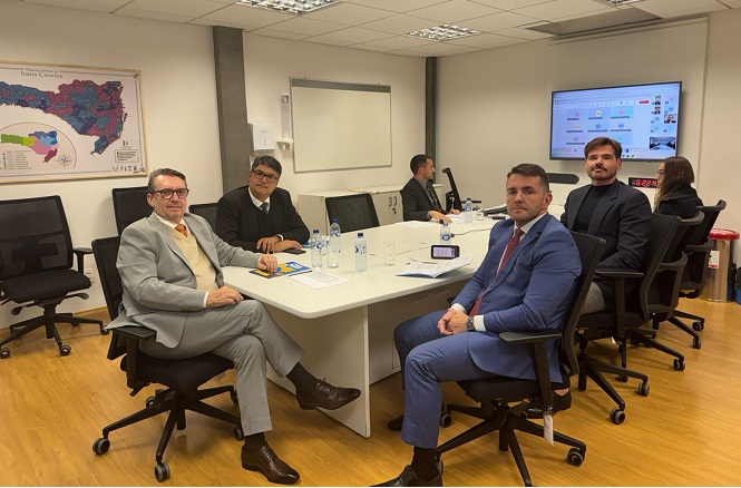 Na imagem, vemos uma sala de reunião onde estão presentes seis pessoas. Quatro delas estão sentadas ao redor de uma mesa de conferência, enquanto as outras duas estão no fundo da sala. As pessoas ao redor da mesa estão vestidas formalmente, usando ternos. Sobre a mesa, há garrafas de água e papéis. Atrás, na parede, há um mapa geográfico emoldurado e uma tela que exibe uma apresentação. A sala parece bem iluminada, com piso de madeira e decoração simples.