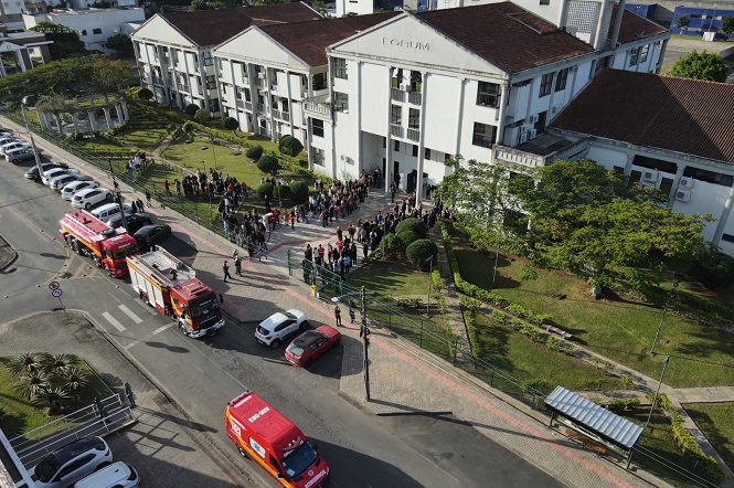 A imagem mostra uma vista aérea de um prédio com a palavra "FÓRUM" na fachada. Em frente ao edifício, há uma concentração de pessoas formando uma fila na calçada, sugerindo que estão aguardando para entrar ou deixar o local. Na rua, há veículos de emergência, incluindo caminhões vermelhos de bombeiros e uma ambulância. A área ao redor tem gramado e árvores, e há uma calçada que segue ao longo da rua, onde outros veículos estão estacionados. O cenário ocorre durante o dia, com boa iluminação natural.