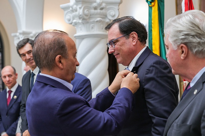 A imagem mostra um grupo de homens em um ambiente formal. No centro, um homem está colocando uma medalha ou insígnia no paletó de outro homem. Ambos estão usando ternos. Ao fundo, há outras pessoas assistindo à cena. O local possui uma decoração elegante com colunas e há bandeiras visíveis à direita. Parece ser uma cerimônia ou evento oficial.