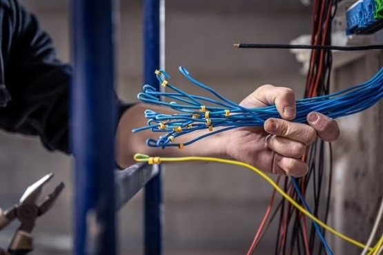 A imagem mostra uma mão segurando vários cabos azuis com conectores nas pontas. Ao redor, há fios de outras cores, como vermelho e preto. À esquerda, pode-se ver uma ferramenta de corte de cabos. A cena parece ser de uma instalação elétrica ou de cabeamento.