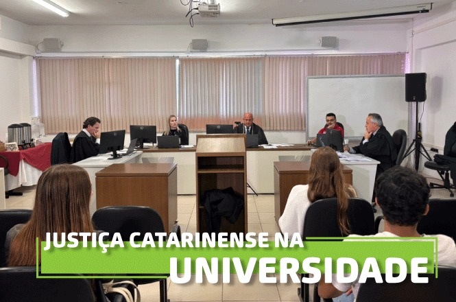 A imagem mostra uma sala de audiências onde está ocorrendo um julgamento simulado ou uma apresentação judicial. Várias pessoas estão sentadas atrás de mesas, possivelmente estudantes e professores ou profissionais da área jurídica. Ao fundo, uma mesa principal com três pessoas indica onde estão os juízes ou mediadores. O ambiente tem uma aparência formal e acadêmica. Na parte inferior, há um texto sobreposto que diz "Justiça Catarinense na Universidade".