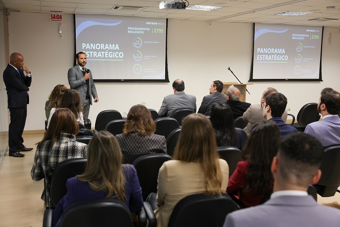 A imagem mostra uma apresentação em um ambiente corporativo. Um homem está de pé, segurando um microfone enquanto fala para a plateia. No fundo, há dois telões projetando o título "Panorama Estratégico" e alguns gráficos, com o texto "Procedimentos Realizados: 2795". A plateia está sentada em cadeiras, assistindo à apresentação. O ambiente parece ser uma sala de conferências ou auditório, com teto embutido e iluminação adequada.