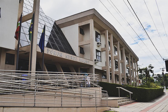 A imagem mostra a fachada de um edifício identificado como "Fórum Nereu Ramos". O prédio é moderno, com uma entrada adornada por bandeiras e um design arquitetônico que incorpora vidro e concreto. Há corrimãos metálicos próximos à entrada, que também possuem escadas. Vê-se parte de uma calçada e a rua ao lado do edifício. O tempo está nublado, com fios elétricos visíveis no céu.