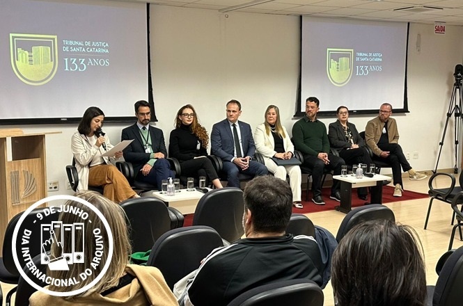 A imagem mostra um grupo de pessoas sentadas em uma mesa redonda em um auditório. Ao fundo, há um telão exibindo o logotipo do Tribunal de Justiça de Santa Catarina, indicando seus 133 anos. As pessoas aparentam estar participando de um evento ou discussão. No canto inferior esquerdo, há um selo com a inscrição "9 de Junho, Dia Internacional do Arquivo". Algumas pessoas na plateia estão de costas, assistindo à apresentação.