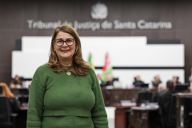 Na imagem, há uma mulher de óculos e cabelos castanhos, vestindo um suéter verde, sorrindo para a câmera. Ela está em um ambiente interno, aparentando ser um tribunal. Ao fundo, é possível ver pessoas sentadas e um letreiro que diz "Tribunal de Justiça de Santa Catarina". Há também bandeiras no ambiente.