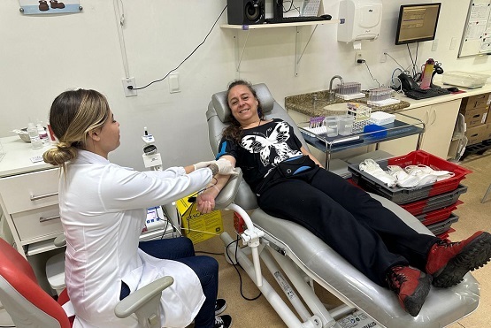 A imagem mostra uma pessoa deitada em uma cadeira de doação de sangue, enquanto outra pessoa, vestida com jaleco branco, parece estar realizando o procedimento de coleta de sangue. A pessoa na cadeira está sorridente e parece à vontade. O ambiente é limpo e organizado, com equipamentos médicos e suprimentos visíveis ao fundo, indicando que é uma clínica ou centro de saúde. Uma bancada ao lado tem bandejas e recipientes usados no processo de doação.
