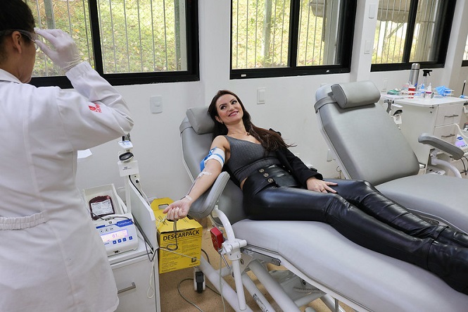 A imagem mostra uma mulher deitada em uma cadeira de doação de sangue, aparentemente em um ambiente de saúde, como um hospital ou clínica. Ela está com um sorriso no rosto enquanto uma profissional de jaleco branco, à esquerda, parece estar assistindo ou auxiliando no processo de doação. Há equipamentos médicos próximos, incluindo uma máquina que parece estar coletando sangue e uma caixa amarela de descarte para objetos cortantes. O ambiente é bem iluminado, com janelas ao fundo permitindo a entrada de luz natural.