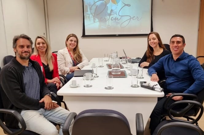 Na imagem, há cinco pessoas sentadas ao redor de uma mesa de reunião em uma sala de conferências. A mesa está coberta com alguns copos, xícaras e um projetor ao fundo exibe a imagem de "PertenSer". As pessoas estão sorrindo e parecem ter uma discussão ou reunião amigável. A sala tem um clima profissional e acolhedor.