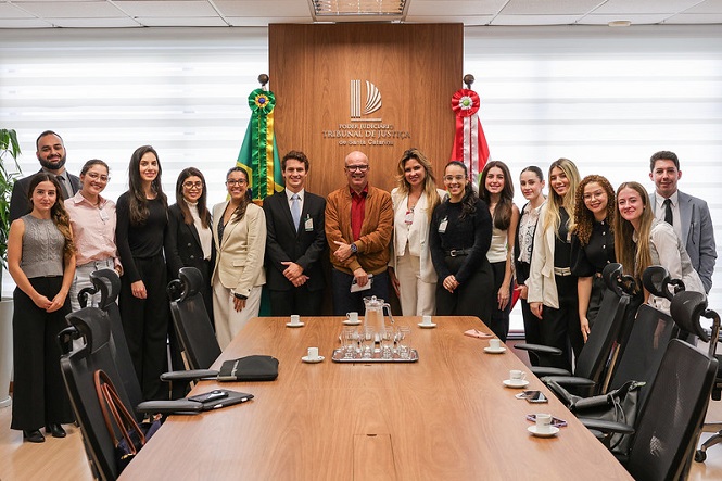 A imagem mostra um grupo de 16 pessoas posando em uma sala de reunião. Eles estão de pé em frente a uma mesa longa, que tem algumas xícaras de café, utensílios e pastas. Ao fundo, há uma parede de madeira com um logotipo e as palavras "Poder Judiciário Tribunal de Justiça do Distrito Federal e Territórios". À esquerda, há uma bandeira do Brasil e, à direita, uma bandeira que parece ser uma representação judicial. As pessoas estão vestidas formalmente, sugerindo um contexto profissional ou oficial. A expressão delas é de satisfação ou alegria.