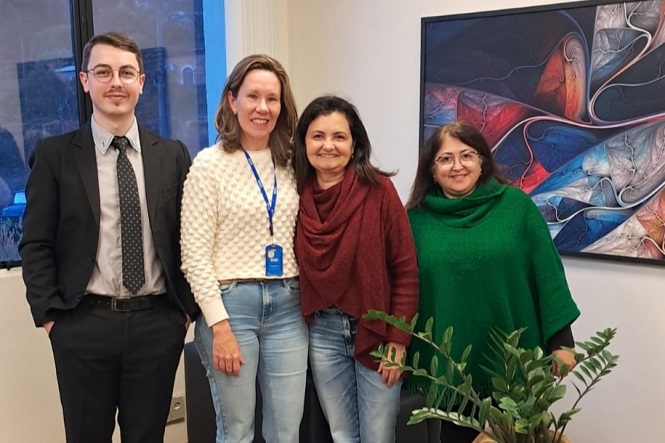 A imagem mostra quatro pessoas posando juntas em um ambiente interno. À esquerda, uma pessoa está vestida com um terno preto e gravata. Ao lado dela, uma segunda pessoa usa um suéter branco e um crachá azul. A terceira pessoa veste um cachecol ou xale vermelho e jeans, enquanto a quarta pessoa está usando um poncho verde. Atrás do grupo há uma pintura abstrata na parede, exibindo uma combinação de cores vibrantes. Também há uma planta no canto inferior direito da imagem. As pessoas estão sorrindo e parecem estar em um ambiente profissional ou de trabalho.