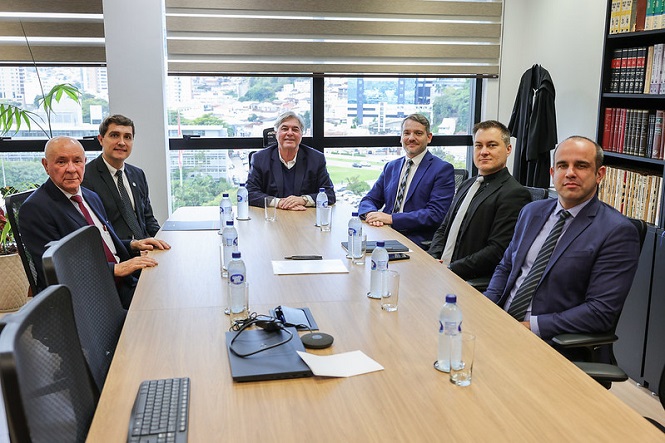 A imagem mostra seis homens sentados em torno de uma mesa de reunião em um escritório. Eles estão vestidos formalmente com ternos. Na mesa há garrafas de água e alguns papéis. Ao fundo, há uma grande janela com vista para a cidade. Nas laterais da sala, podem-se ver estantes com livros.