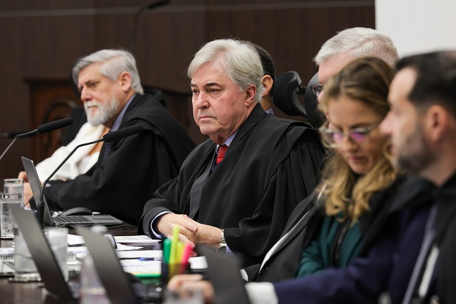 A imagem mostra várias pessoas sentadas lado a lado em uma mesa, provavelmente em um ambiente formal ou oficial, como um tribunal ou reunião importante. Eles estão vestindo trajes formais, alguns com togas pretas. Há laptops na mesa e copos de água. A expressão é séria, indicando que estão participando de uma discussão ou julgamento. A cena parece retratar uma reunião ou sessão judicial.