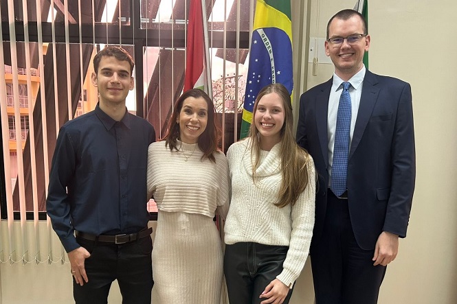A imagem mostra quatro pessoas posando para uma foto lado a lado em um ambiente fechado. Ao fundo, há uma janela com persianas e três bandeiras, sendo uma delas a do Brasil. As pessoas estão vestidas de maneira formal e sorriem para a câmera.