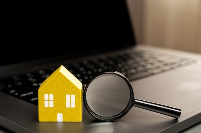 A imagem mostra um modelo de casa em miniatura de cor amarela ao lado de uma lupa. Ambos estão posicionados sobre o teclado de um laptop. A combinação dos elementos sugere uma relação com pesquisa ou busca por imóveis online.