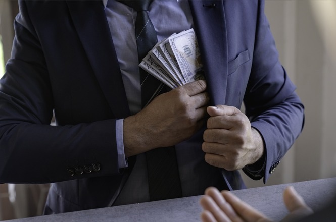 A imagem mostra uma pessoa vestindo um terno azul escuro, colocando várias notas de dinheiro no bolso interno do paletó. A mão esquerda segura as notas, enquanto a direita segura o terno. Em primeiro plano, há uma mesa e as mãos de outra pessoa estão parcialmente visíveis, sugerindo uma interação, possivelmente um gesto de conversa ou negociação.