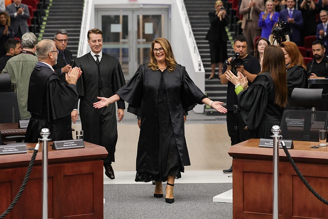 A imagem mostra uma cena em um ambiente formal, provavelmente dentro de um tribunal ou um auditório oficial. No centro, uma mulher usando uma toga preta, parece estar sendo recebida com aplausos. Outras pessoas ao redor também estão usando togas pretas. Elas parecem estar participando de algum evento ou cerimônia oficial. A atmosfera é de celebração ou reconhecimento. Ao fundo, há escadas e mais pessoas assistindo à cena.