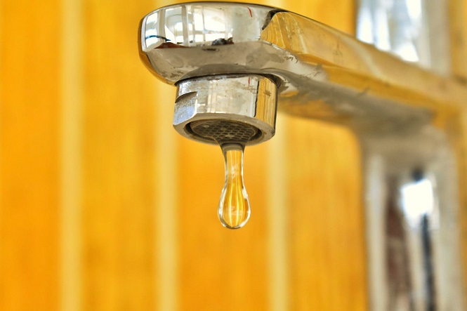 A imagem mostra uma torneira de metal com uma gota de água prestes a cair. O fundo é desfocado e tem uma tonalidade amarelada.