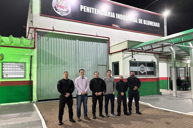 A imagem mostra um grupo de seis pessoas posicionadas na frente da entrada de um edifício. Acima, uma placa identifica o local como "Penitenciária Industrial de Blumenau". As pessoas estão em pé em uma área pavimentada, e a fachada do prédio é pintada com cores claras e detalhes em verde. Não posso identificar quem são as pessoas na imagem.