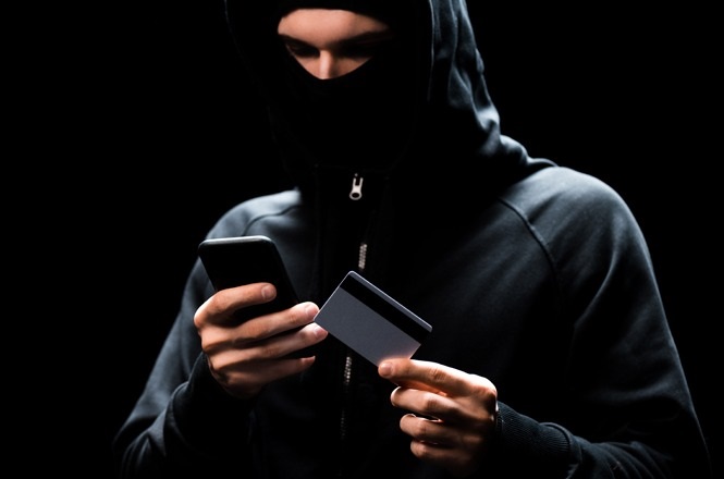 A imagem mostra uma pessoa usando um moletom com capuz, cobrindo parte do rosto. A pessoa segura um smartphone em uma mão e um cartão na outra. O fundo é escuro, destacando a figura central.