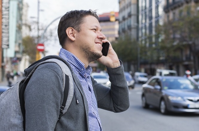 A imagem mostra um homem em pé em uma calçada de uma rua urbana movimentada. Ele está de perfil, segurando um telefone celular ao ouvido com a mão direita, aparentando estar em uma conversa. O homem veste uma camisa xadrez azul e branca por baixo de um casaco cinza e carrega uma mochila cinza nas costas. Ao fundo, há carros passando e edifícios altos, indicando que ele está em uma cidade. Ele parece estar sorrindo ou conversando animadamente.