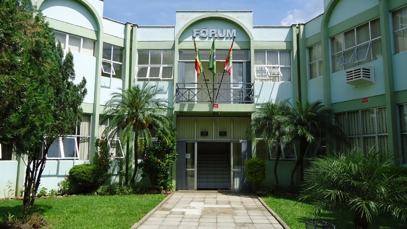 A imagem mostra a fachada de um prédio identificado como "FÓRUM". O edifício possui uma arquitetura simétrica com janelas amplas e portas de vidro na entrada principal. Há três bandeiras hasteadas acima da entrada. O caminho em direção à entrada é pavimentado e ladeado por grama e plantas, incluindo palmeiras. A fachada do prédio tem um tom claro de verde, e o dia está ensolarado.