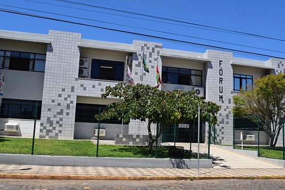 A imagem mostra um prédio com fachada de azulejos cinza e algumas janelas. No lado direito, está escrita a palavra "FÓRUM" em letras grandes. Há uma calçada e um gramado em frente ao edifício, com algumas árvores. Na fachada, há bandeiras hasteadas. É um dia ensolarado com céu azul e algumas linhas de fios elétricos estão visíveis no topo da imagem.