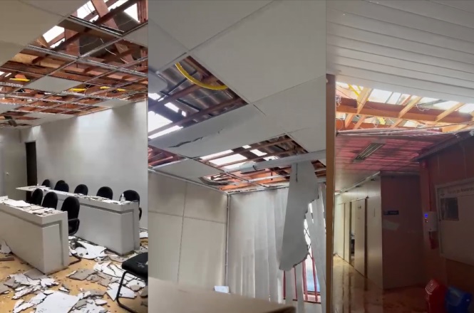 A imagem mostra um ambiente interno com danos significativos no teto. Há placas de forro caídas e algumas áreas expostas revelando a estrutura de madeira e possíveis tubulações. No chão, há escombros espalhados, indicando que partes do teto desabaram. À esquerda, há uma sala com cadeiras, semelhante a uma sala de conferências ou aula. À direita, um corredor também exibe danos no teto, com elementos estruturais visíveis. O ambiente parece desorganizado e danificado.