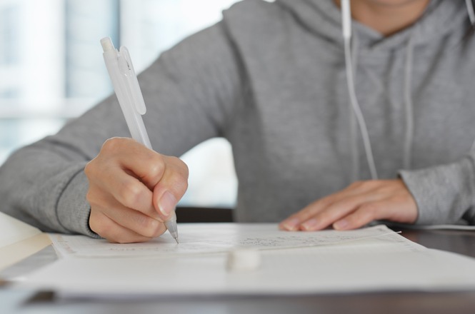 A imagem mostra uma pessoa escrevendo em um caderno ou bloco de notas. Ela está usando uma caneta de aparência branca. A pessoa veste um moletom cinza e tem fones de ouvido brancos. A mesa está desfocada no plano de fundo, e a imagem dá a sensação de estudo ou trabalho.