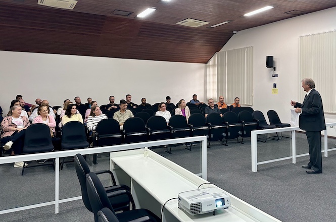 A imagem mostra uma sala de aula ou auditório onde um grupo de pessoas está sentado em cadeiras, prestando atenção em um palestrante que está em pé à frente. O ambiente é iluminado, com teto de madeira e paredes brancas. Há mesas próximas ao palestrante, e um projetor está colocado sobre elas, indicando que pode haver uma apresentação. O palestrante está vestido formalmente e parece estar explicando e discutindo algum tópico com a plateia.