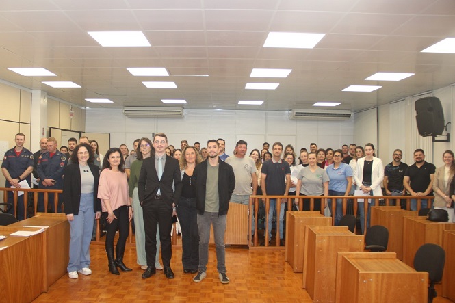 A imagem mostra um grupo grande de pessoas em uma sala. Na frente, há algumas pessoas em destaque, possivelmente autoridades ou palestrantes. Atrás delas está um grupo maior, posicionado em pé. O ambiente parece ser uma sala de conferências ou uma sala de aula, com cadeiras e mesas de madeira ao redor. O teto tem iluminação fluorescente.