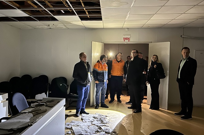 A imagem mostra um grupo de pessoas em um ambiente interno, possivelmente um escritório ou sala de conferências, que aparenta estar danificado. Parte do teto está caído no chão e há sinais de deterioração. Algumas pessoas estão usando roupas casuais e outras estão de uniforme laranja, geralmente associado a equipes de emergência ou segurança. A iluminação é clara sobre o grupo, enquanto o restante da sala está em sombras. Há uma placa de "SAÍDA" acima da porta aberta no fundo.