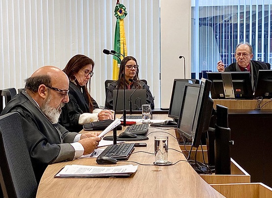 A imagem mostra um grupo de pessoas em um ambiente interno, possivelmente um escritório ou sala de conferências, que aparenta estar danificado. Parte do teto está caído no chão e há sinais de deterioração. Algumas pessoas estão usando roupas casuais e outras estão de uniforme laranja, geralmente associado a equipes de emergência ou segurança. A iluminação é clara sobre o grupo, enquanto o restante da sala está em sombras. Há uma placa de "SAÍDA" acima da porta aberta no fundo.