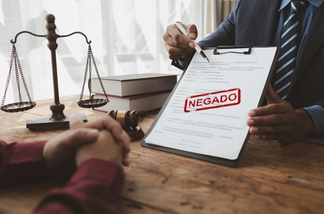 A imagem mostra duas pessoas sentadas em uma mesa. Uma delas está segurando uma prancheta com um documento, que tem a palavra "NEGADO" em destaque. Também há uma balança de justiça e alguns livros sobre a mesa, sugerindo um ambiente ligado a questões jurídicas ou legais.