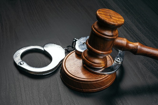 A imagem mostra um martelo de juiz de madeira pousado em cima de uma base. Ao lado do martelo, há um par de algemas de metal. Ambos os objetos estão sobre uma superfície escura. A cena simboliza a justiça e a lei.