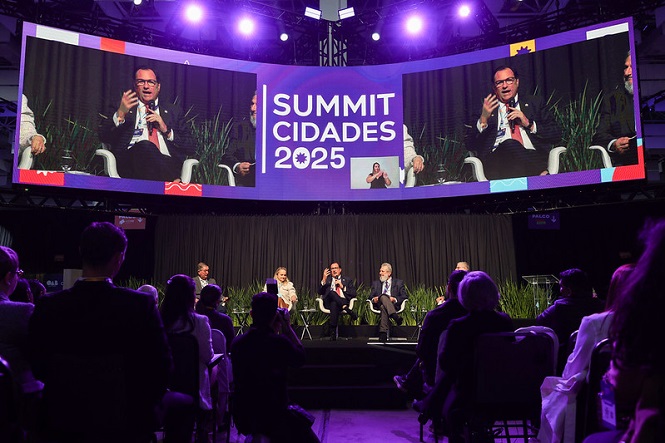 Foto horizontal: no telão, desembargador Francisco Oliveira Neto discursa no Summit Cidades 2025, público e painel ao vivo, tons roxo e azul.