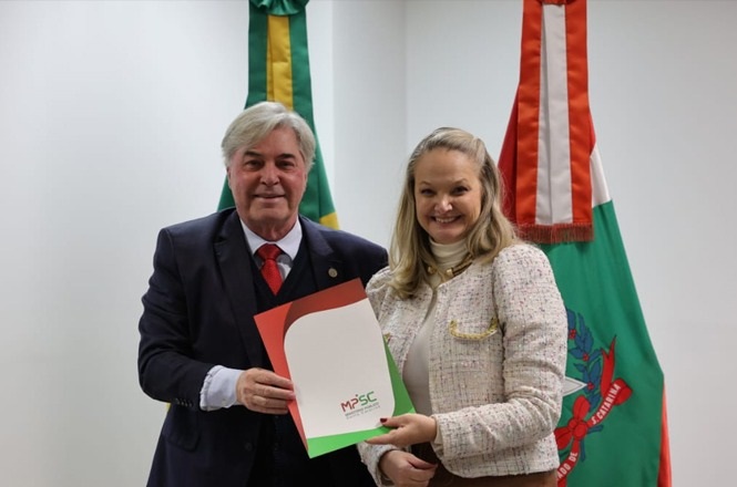 Na imagem, há um homem e uma mulher posando para uma foto. Os dois estão segurando uma pasta que tem o logo do Ministério Público de Santa Catarina (MPSC). Atrás deles, há bandeiras, uma do Brasil e outra de Santa Catarina. As pessoas estão sorrindo e parecem estar em um ambiente formal, provavelmente um escritório ou uma instituição pública.